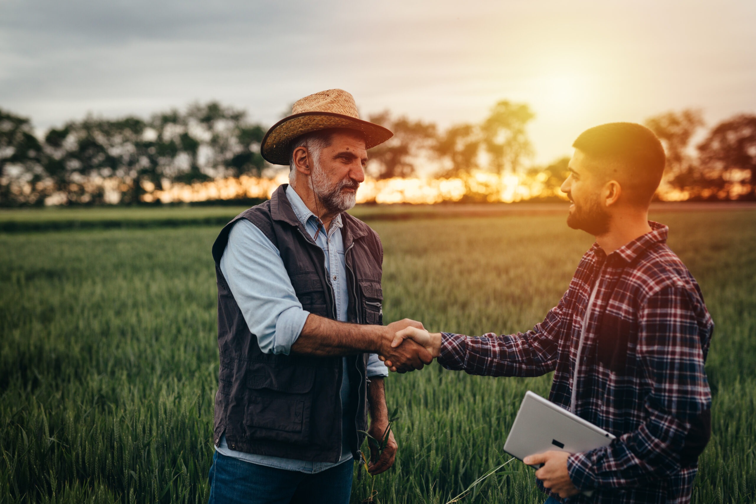 Two men shake hands in a lush green field during sunset. One wears a straw hat, while the other holds a tablet, symbolizing collaboration in agriculture.